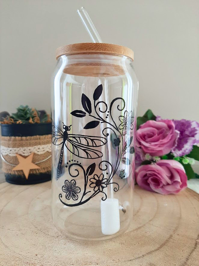 Gobelet paille en verre peint à la main avec libellule et motifs floraux, couvercle en bambou et paille en verre, posé sur une table en bois avec des roses roses