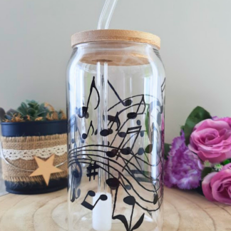 Gobelet paille en verre peint à la main avec partition musicale et notes virevoltantes noires, couvercle en bambou et paille en verre, posé sur une table en bois avec des roses roses
