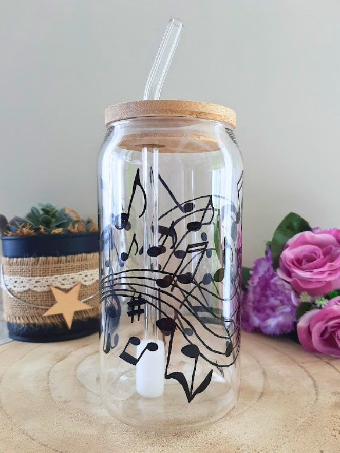 Gobelet paille en verre peint à la main avec partition musicale et notes virevoltantes noires, couvercle en bambou et paille en verre, posé sur une table en bois avec des roses roses