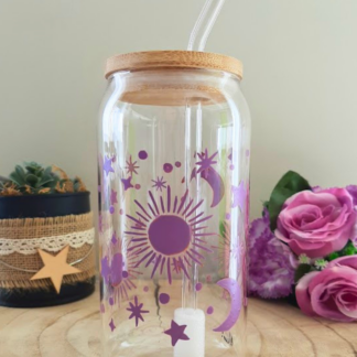 Gobelet paille en verre peint à la main avec soleil lune et étoiles violet, couvercle en bambou et paille en verre, posé sur une table en bois avec des roses violettes