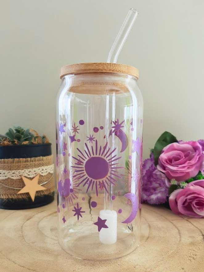 Gobelet paille en verre peint à la main avec soleil lune et étoiles violet, couvercle en bambou et paille en verre, posé sur une table en bois avec des roses violettes