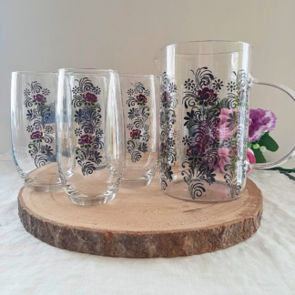 Set pichet et 4 verres en verre peints à la main avec motif floral folklorique roses bordeaux et noir, posés sur une rondelle de bois avec des fleurs roses en arrière-plan