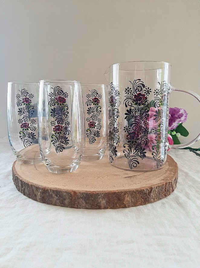 Set pichet et 4 verres en verre peints à la main avec motif floral folklorique roses bordeaux et noir, posés sur une rondelle de bois avec des fleurs roses en arrière-plan