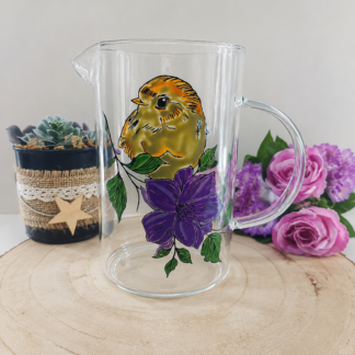 Pichet en verre peint à la main avec oisillon jaune et fleur violette, posé sur une table en bois avec des roses roses