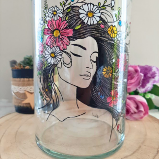 Vase en verre 20cm peint à la main avec portrait féminin aux yeux clos couronné de fleurs multicolores, posé sur une table en bois avec des roses violettes