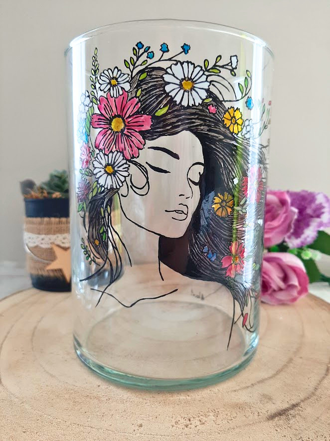 Vase en verre 20cm peint à la main avec portrait féminin aux yeux clos couronné de fleurs multicolores, posé sur une table en bois avec des roses violettes