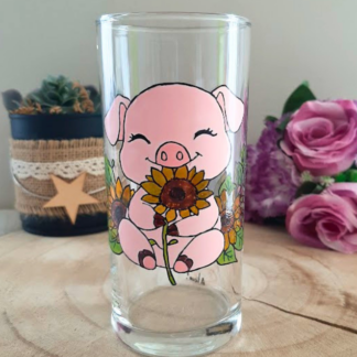 Verre droit peint à la main avec petit cochon rose tenant un tournesol, posé sur une table en bois avec des roses roses