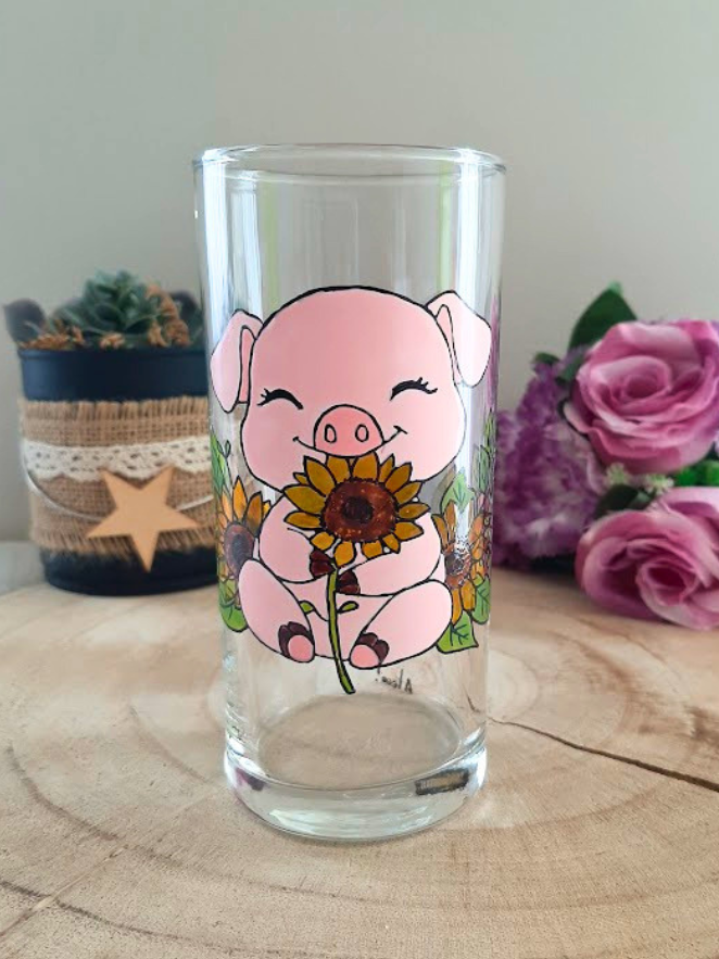 Verre droit peint à la main avec petit cochon rose tenant un tournesol, posé sur une table en bois avec des roses roses