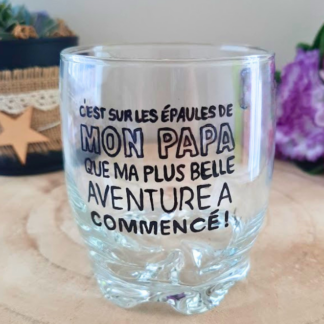 Verre à whisky peint à la main avec inscription "C'est sur les épaules de mon papa que ma plus belle aventure a commencé", posé sur une table en bois avec des roses roses