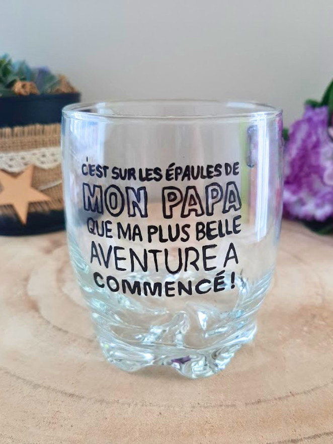 Verre à whisky peint à la main avec inscription "C'est sur les épaules de mon papa que ma plus belle aventure a commencé", posé sur une table en bois avec des roses roses