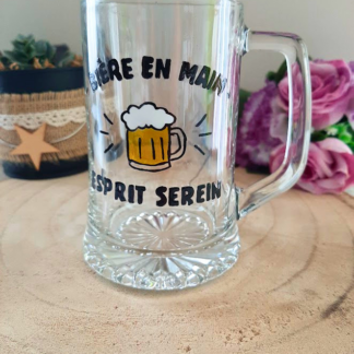 Chope à bière peinte à la main avec inscription "Bière en main, esprit serein" et chope de bière mousseuse colorée, posée sur une table en bois avec des roses roses
