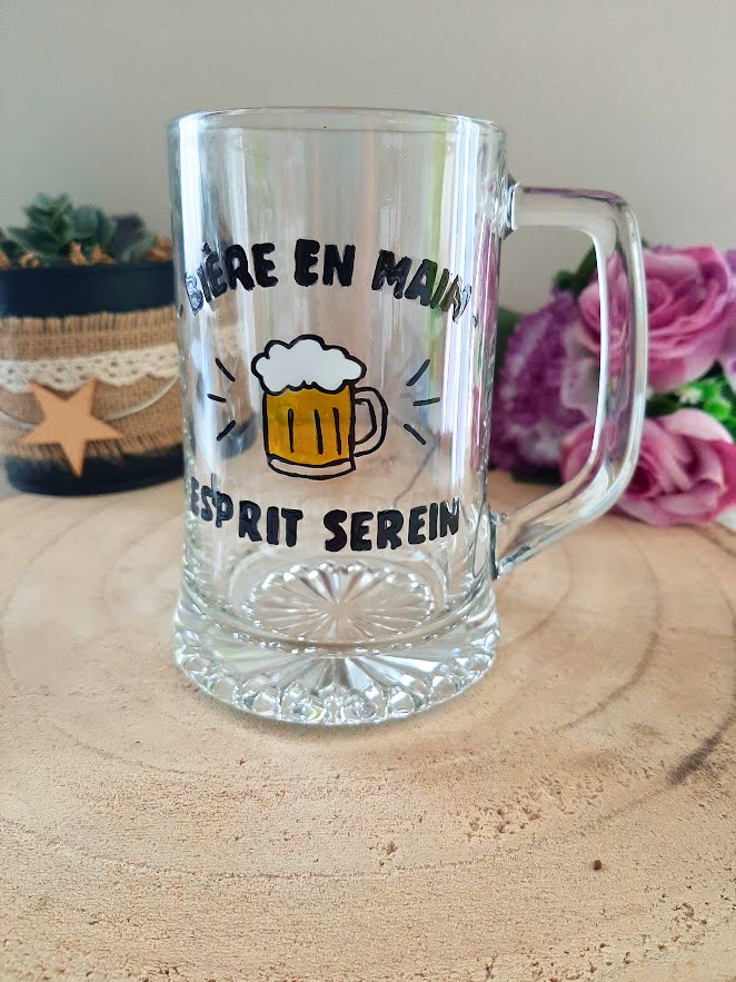 Chope à bière peinte à la main avec inscription "Bière en main, esprit serein" et chope de bière mousseuse colorée, posée sur une table en bois avec des roses roses