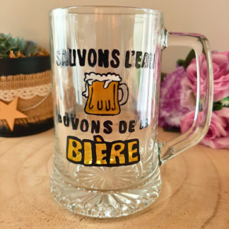Chope à bière peinte à la main avec inscription "Sauvons l'eau, buvons de la bière" en noir et jaune, posée sur une table en bois avec des roses roses