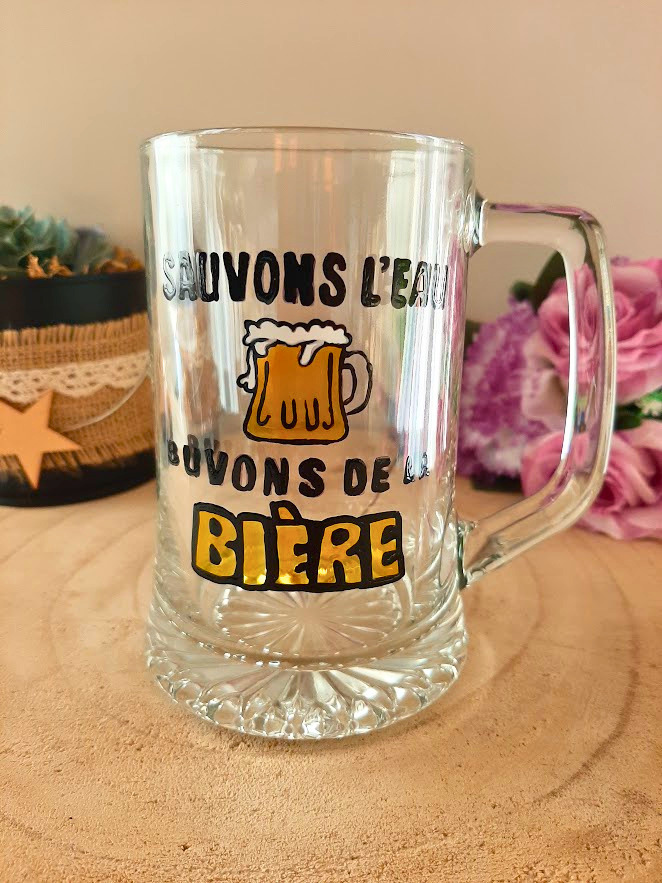 Chope à bière peinte à la main avec inscription "Sauvons l'eau, buvons de la bière" en noir et jaune, posée sur une table en bois avec des roses roses