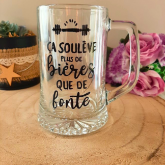 Chope à bière peinte à la main avec inscription "Ça soulève plus de bières que de fonte" et haltère, posée sur une table en bois avec des roses roses
