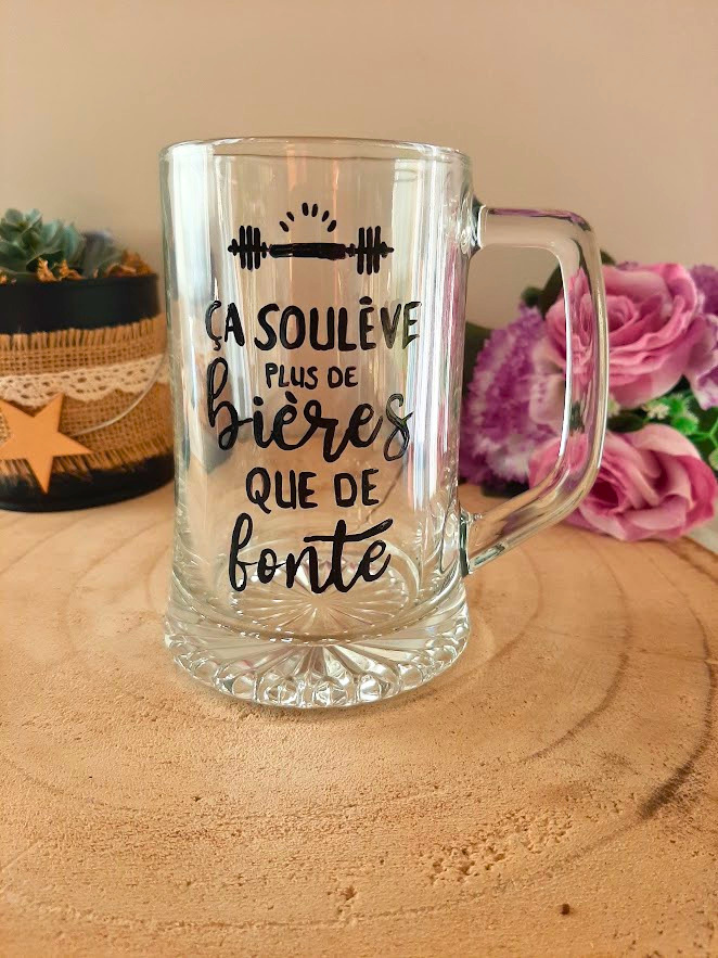 Chope à bière peinte à la main avec inscription "Ça soulève plus de bières que de fonte" et haltère, posée sur une table en bois avec des roses roses