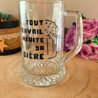 Chope à bière peinte à la main avec inscription "Tout travail mérite sa bière" et chope mousseuse dessinée, posée sur une table en bois avec des roses roses