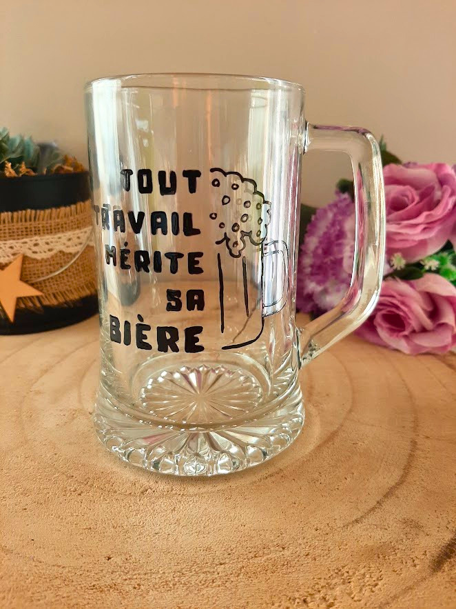 Chope à bière peinte à la main avec inscription "Tout travail mérite sa bière" et chope mousseuse dessinée, posée sur une table en bois avec des roses roses