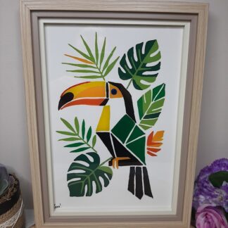Tableau sur verre peint à la main avec toucan géométrique et feuillage tropical, encadré en bois naturel format A4, posé sur une table en bois avec des roses roses