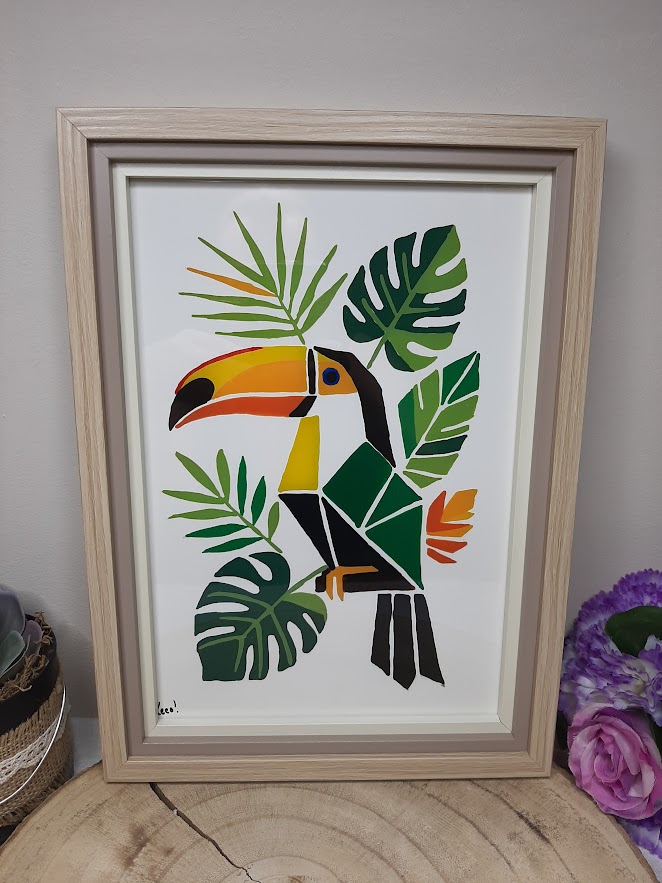Tableau sur verre peint à la main avec toucan géométrique et feuillage tropical, encadré en bois naturel format A4, posé sur une table en bois avec des roses roses