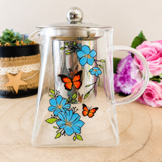 Théière en verre peinte à la main avec fleurs bleues et papillons orangés, couvercle acier inoxydable, posée sur une table en bois avec des roses roses