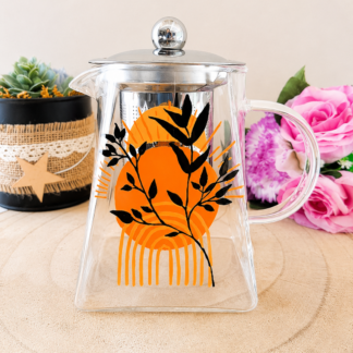 Théière en verre peinte à la main avec grand soleil orange style boho et feuillage noir, couvercle acier inoxydable, posée sur une table en bois avec des roses roses