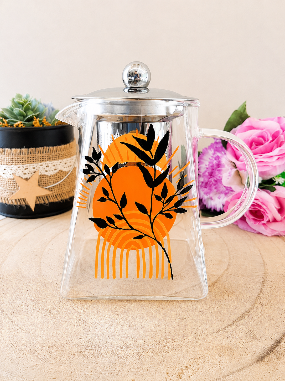 Théière en verre peinte à la main avec grand soleil orange style boho et feuillage noir, couvercle acier inoxydable, posée sur une table en bois avec des roses roses