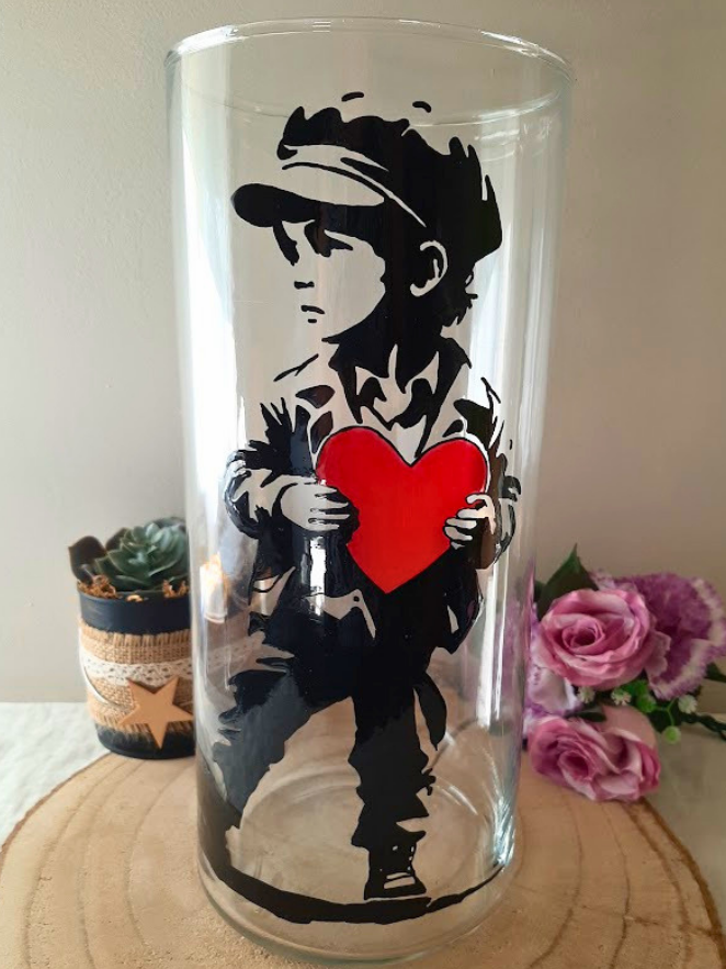 Grand vase cylindrique en verre peint à la main avec enfant style street art tenant un cœur rouge, posé sur une table en bois avec des roses roses