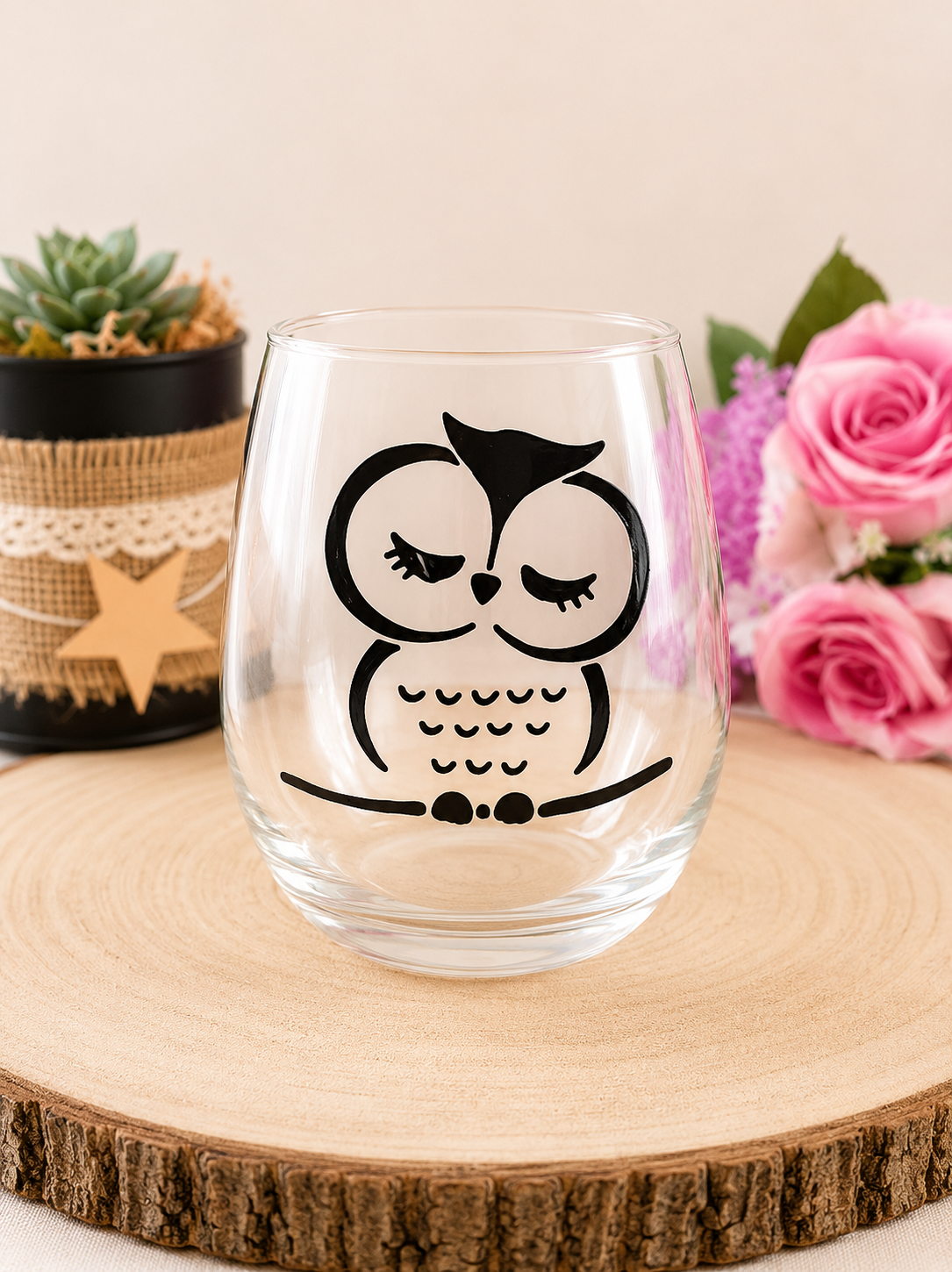 Gobelet en verre peint à la main avec hibou endormi noir et blanc style graphique épuré, posé sur une table en bois avec des roses roses