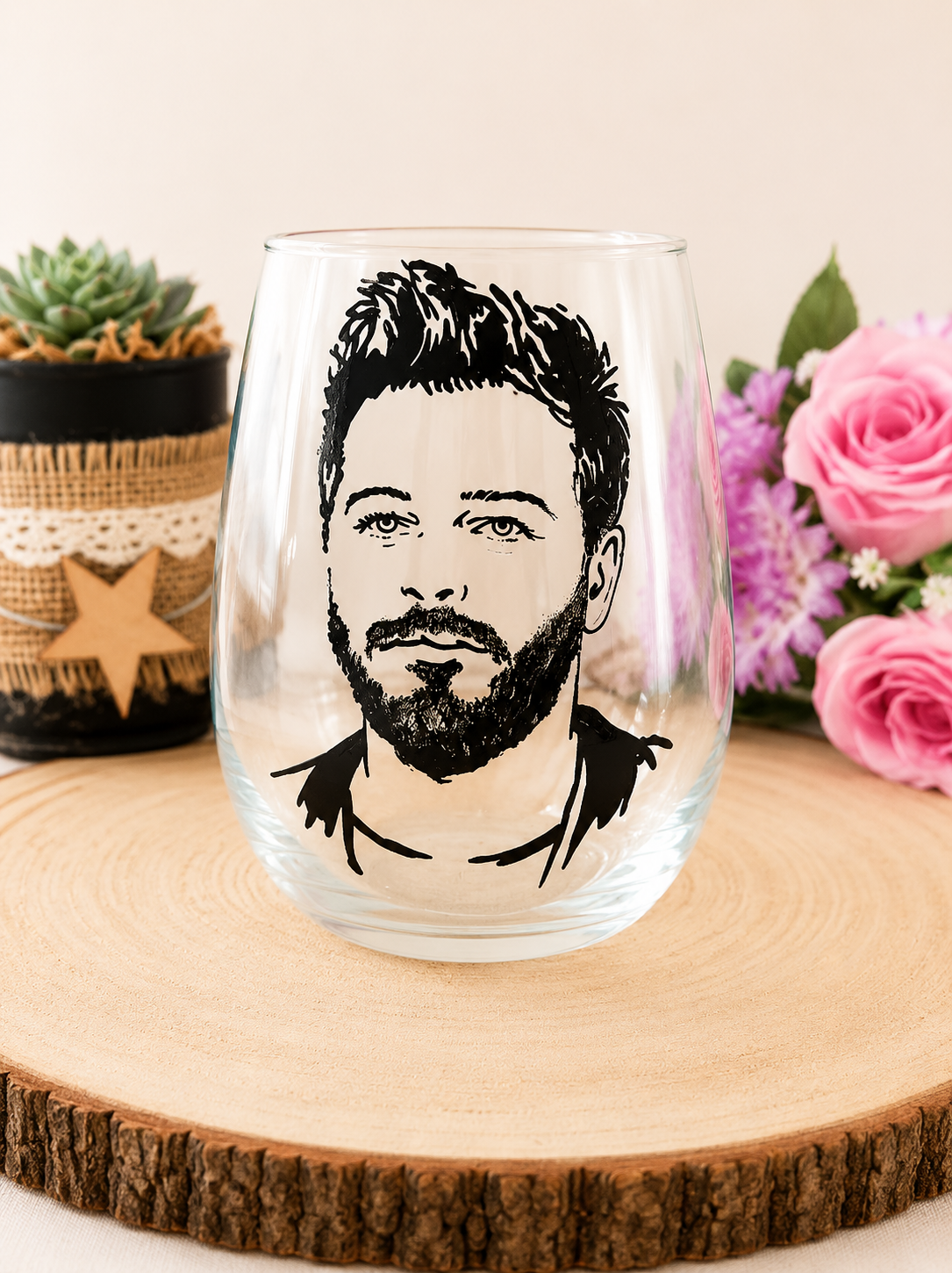 Gobelet en verre peint à la main avec portrait réaliste de Christophe Maé en noir et blanc, posé sur une table en bois avec des roses roses