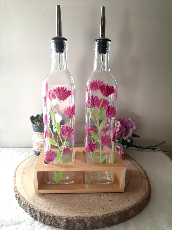 Duo huilier et vinaigrier en verre peints à la main avec grandes fleurs roses et feuillage vert, présentés dans leur support en bois naturel sur une table en bois