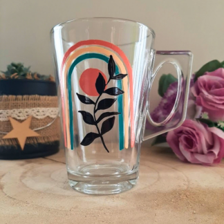 Mug en verre peint à la main avec arc-en-ciel pastel et feuillage noir, posé sur une table en bois