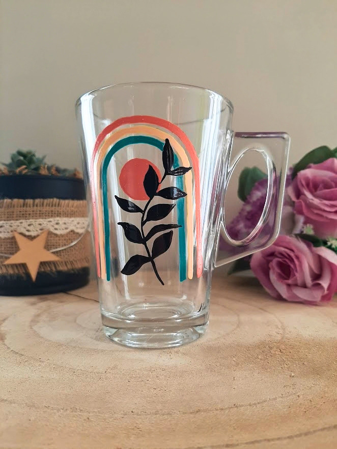 Mug en verre peint à la main avec arc-en-ciel pastel et feuillage noir, posé sur une table en bois