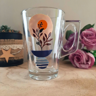 Mug en verre peint à la main avec vase bleu, bouquet bordeaux et soleil orange, posé sur une table en bois