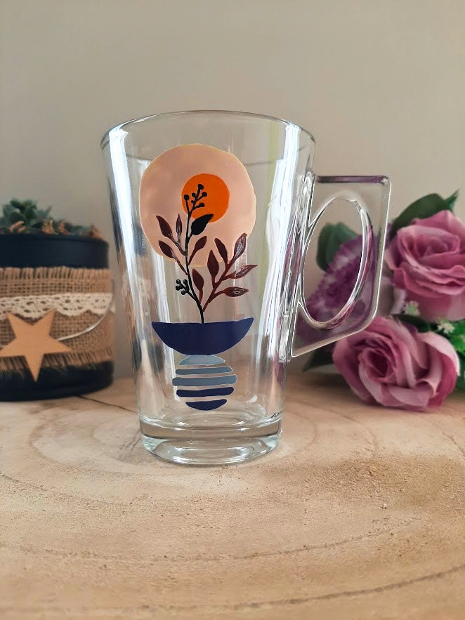 Mug en verre peint à la main avec vase bleu, bouquet bordeaux et soleil orange, posé sur une table en bois