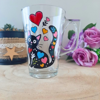 Mug en verre peint à la main avec chat noir et cœurs multicolores, posé sur une table en bois