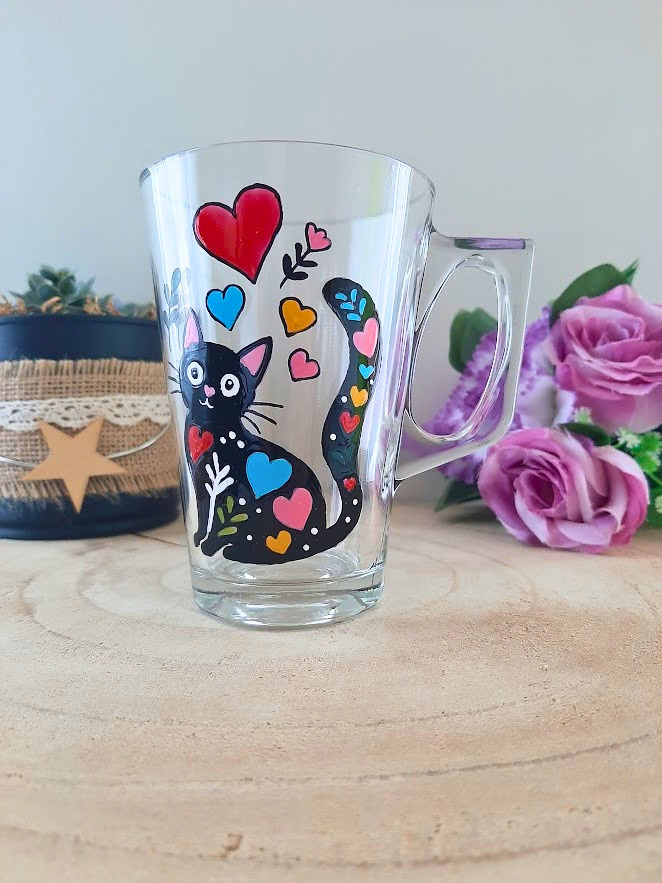 Mug en verre peint à la main avec chat noir et cœurs multicolores, posé sur une table en bois