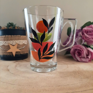 Mug en verre peint à la main avec formes géométriques orange, vert et feuillage noir, posé sur une table en bois
