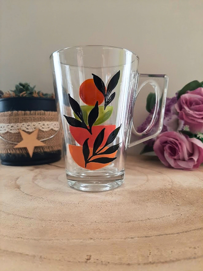 Mug en verre peint à la main avec formes géométriques orange, vert et feuillage noir, posé sur une table en bois