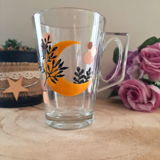 Mug en verre peint à la main avec croissant de lune orange et feuillage, posé sur une table en bois