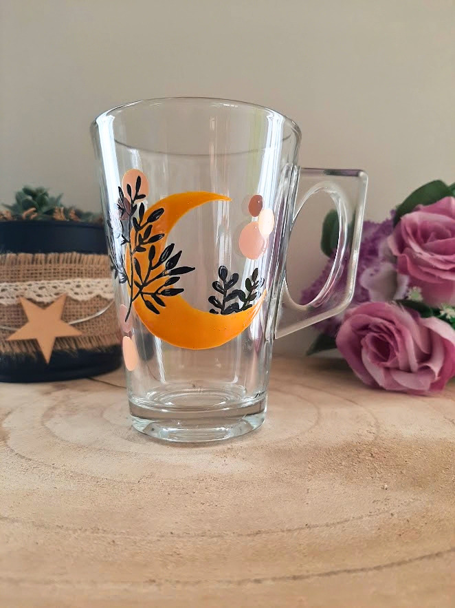 Mug en verre peint à la main avec croissant de lune orange et feuillage, posé sur une table en bois