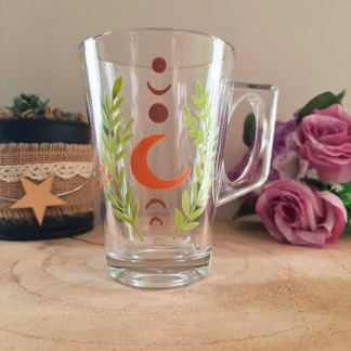 Mug en verre peint à la main avec phases de lune et feuillage vert, posé sur une table en bois