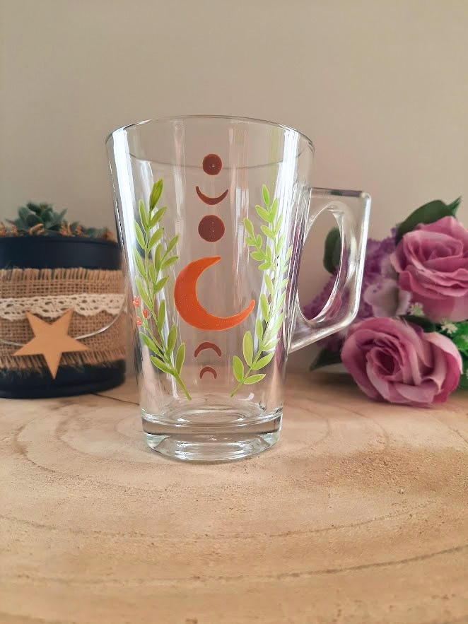 Mug en verre peint à la main avec phases de lune et feuillage vert, posé sur une table en bois