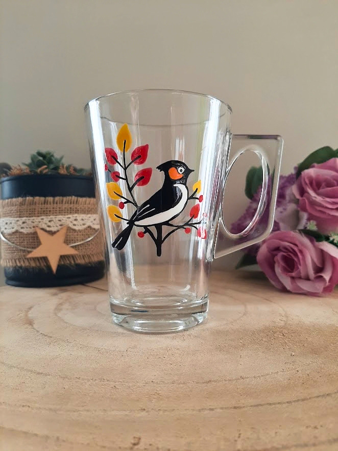 Mug en verre peint à la main avec oiseau sur une branche et feuilles d'automne, posé sur une table en bois