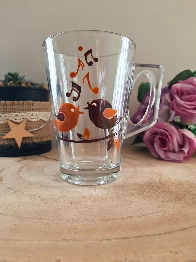 Mug en verre peint à la main avec deux oiseaux et notes de musique, posé sur une table en bois