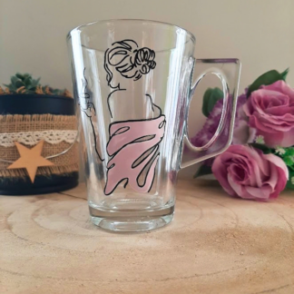 Mug en verre peint à la main avec silhouette de femme et robe rose, posé sur une table en bois avec des roses roses