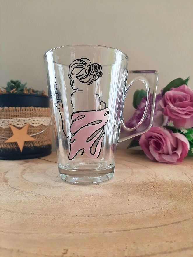 Mug en verre peint à la main avec silhouette de femme et robe rose, posé sur une table en bois avec des roses roses