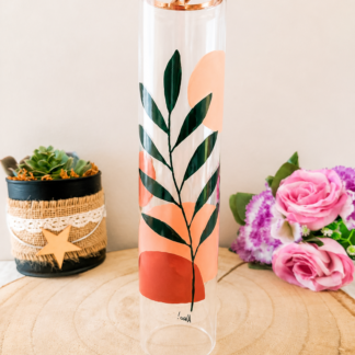 Porte-encens cylindrique en verre peint à la main avec branche de feuillage vert foncé et formes géométriques terracotta et pêche, cerclage doré, posé sur une table en bois avec des roses roses