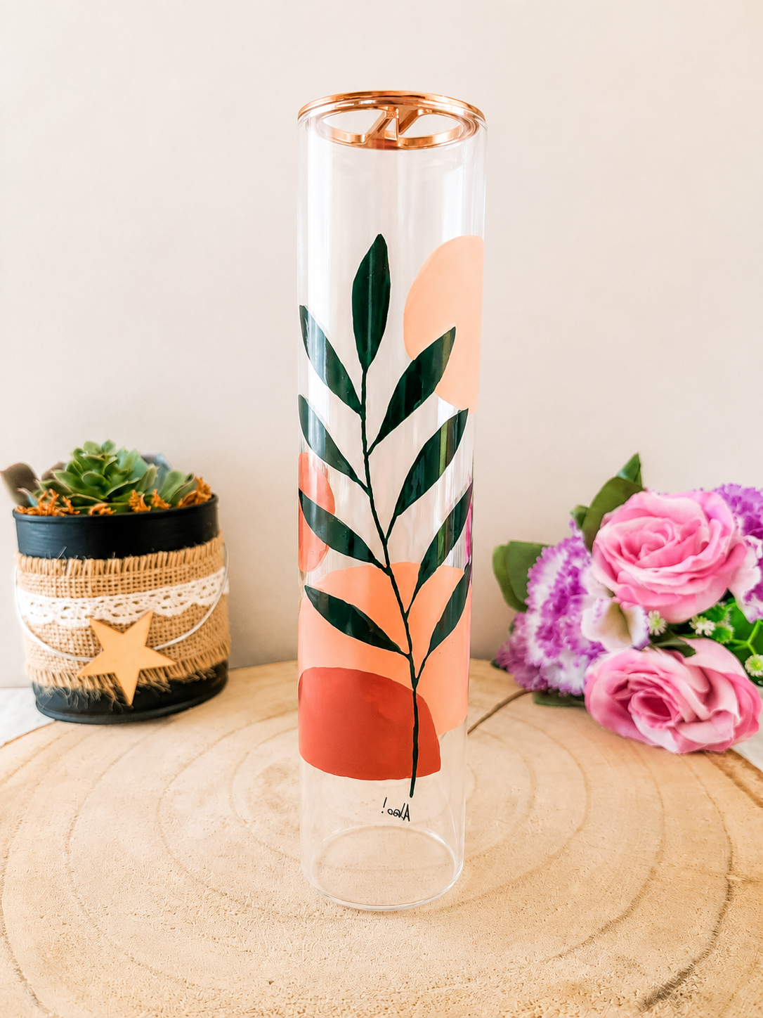 Porte-encens cylindrique en verre peint à la main avec branche de feuillage vert foncé et formes géométriques terracotta et pêche, cerclage doré, posé sur une table en bois avec des roses roses