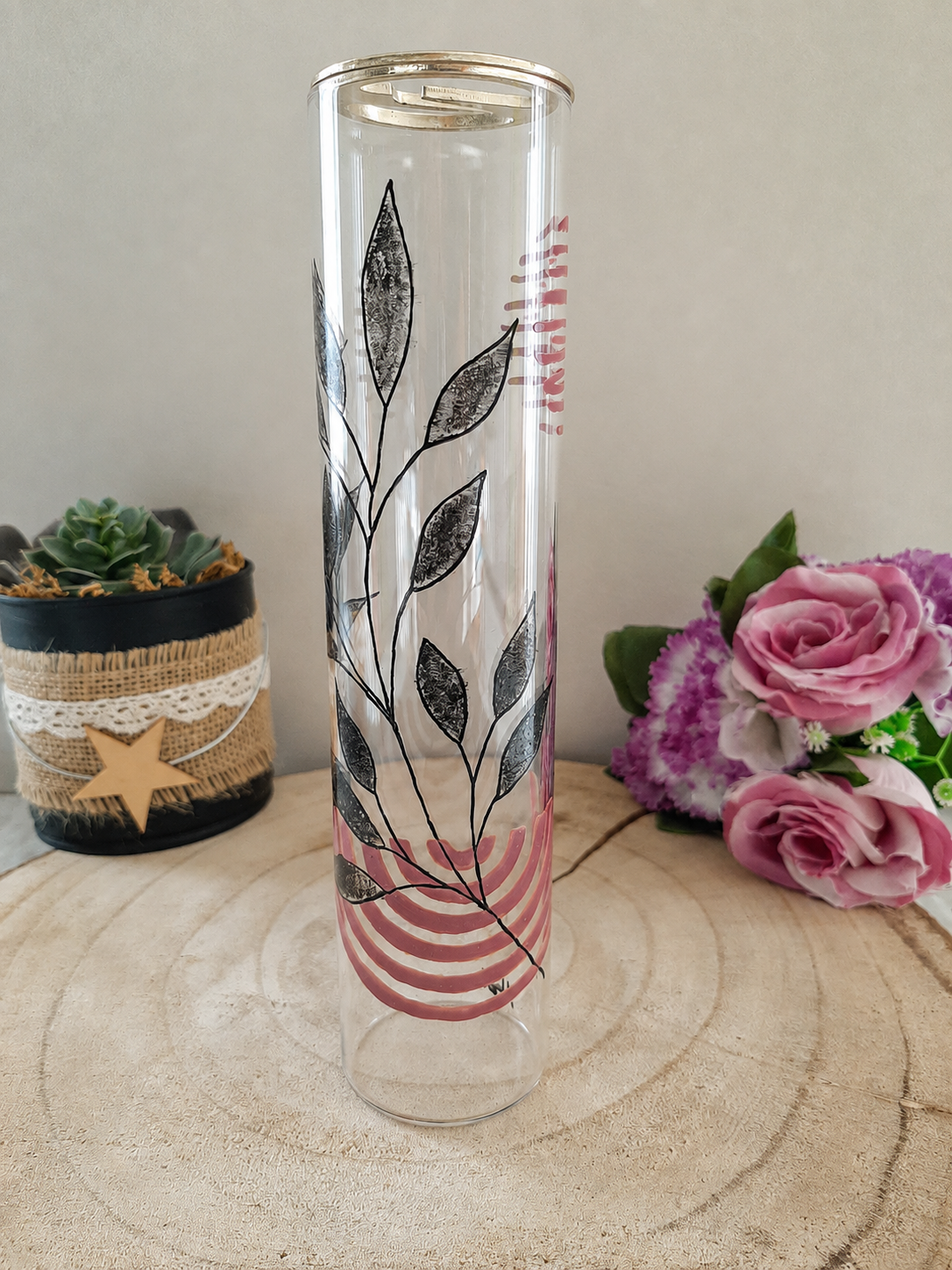 Porte-encens cylindrique en verre peint à la main avec feuillage graphique noir et arcs roses, cerclage argenté, posé sur une table en bois avec des roses roses